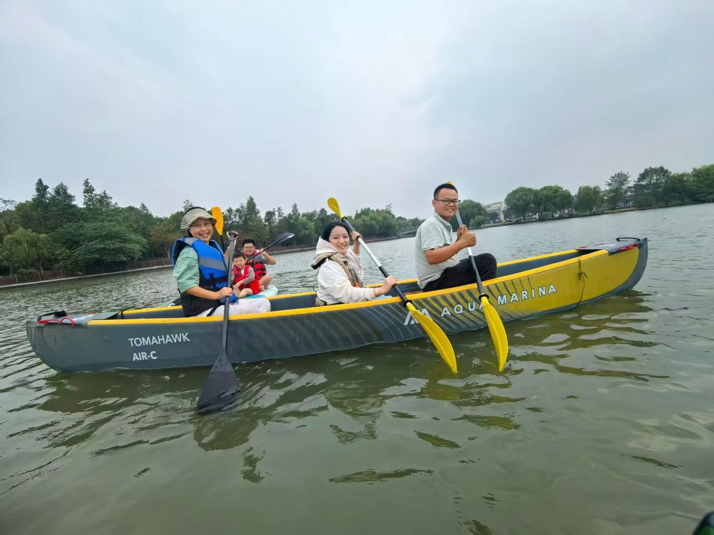 一条黄色双体独木舟上坐着四个人，都穿着救生衣，正在湖面上划桨